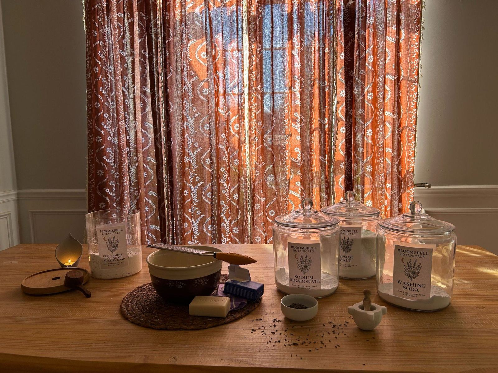 Bloomspell botanicals table — glass jars of sodium bicarbonate and washing soda, mortar and pestle, bar soap, candle, and scattered lavender buds on a wooden table.